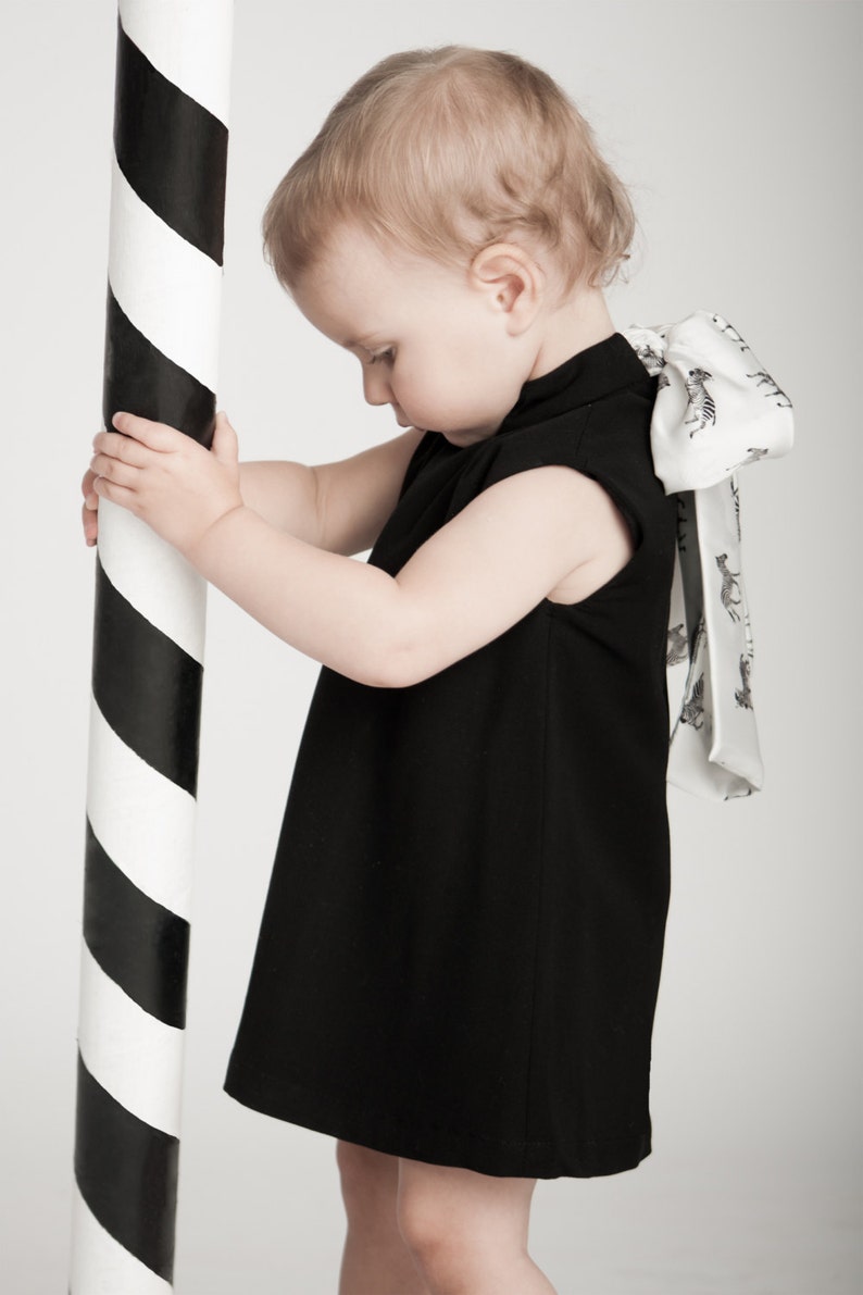 May include: A young child wearing a black sleeveless dress with a white bow tied at the back. The bow has a zebra print pattern. The child is holding onto a black and white striped pole.