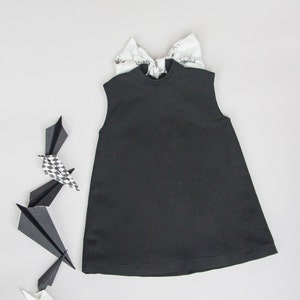 May include: A black sleeveless dress with a high neck and a bow on the back. The dress is laying flat on a white surface with paper airplanes around it.