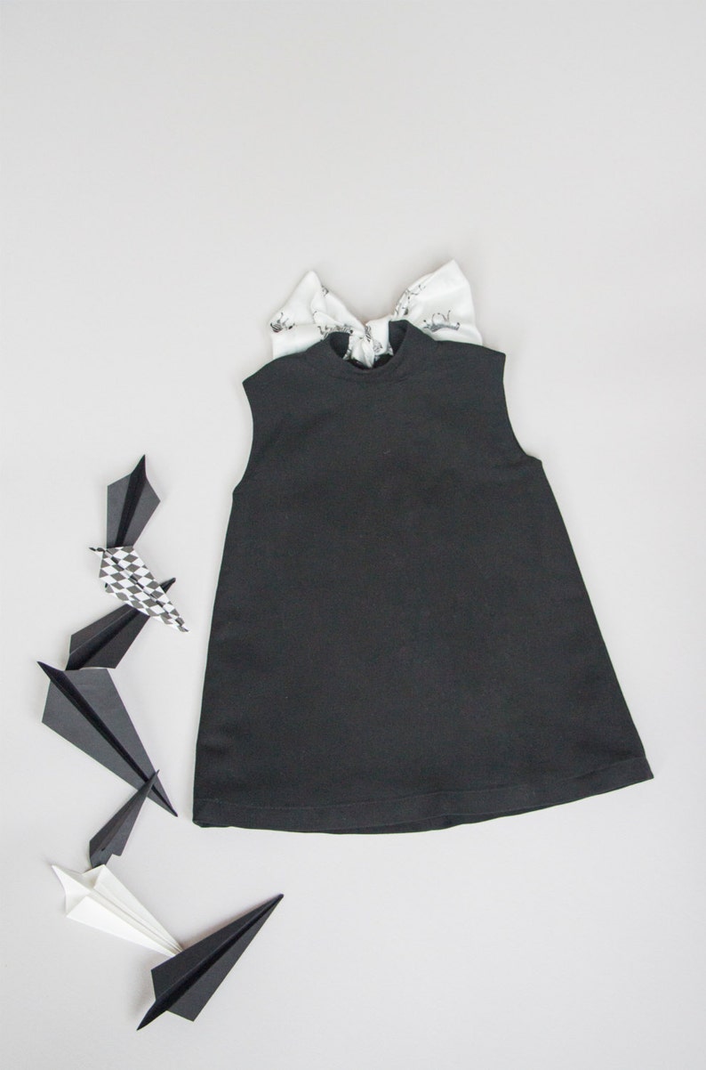 May include: A black sleeveless dress with a high neck and a bow on the back. The dress is laying flat on a white surface with paper airplanes around it.