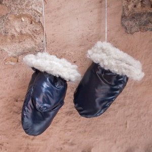 May include: Two navy blue mittens with a shiny finish hang against a textured, light brown wall. Each mitten has a fluffy, off-white cuff. The mittens are suspended by thin, silver-colored strings.