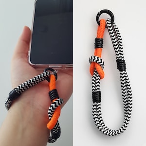 以下が含まれることがあります： オレンジのアクセントが付いた黒と白の編組電話ストラップ。ストラップの上部に黒い金属製のリングがあり、電話に取り付けられています。