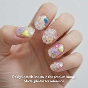 May include: Close-up of a hand with decorated nails. The nails feature a clear base with glitter and various embellishments, including small flowers, hearts, and beads in pastel colors. The text at the bottom reads: Design details shown in the product image. Model photos for reference.
