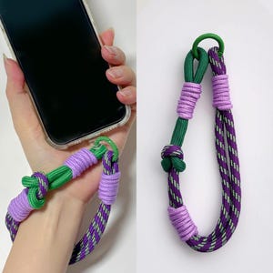 May include: A green and purple phone strap with a green metal ring. The strap is made of braided rope and has a knot at the top.