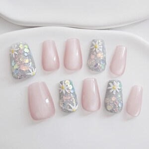 May include: A set of ten artificial nails in a variety of designs. Some nails are a pale pink, while others are covered in silver glitter, sequins, and small white daisy accents. The nails are arranged on a white surface.