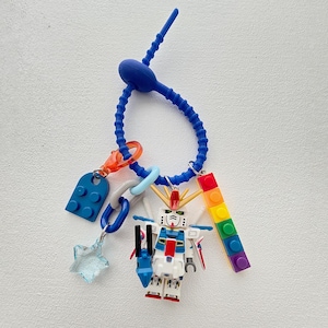 Peut inclure: Un porte-clés en silicone bleu avec divers breloques. Les breloques comprennent un robot jouet, une brique Lego bleue, une brique Lego arc-en-ciel, une étoile transparente et un anneau bleu et orange. Le porte-clés est sur une surface blanche.