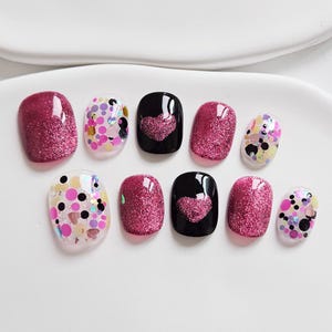 May include: A set of ten artificial nails in various designs. The nails feature glitter, confetti, and heart patterns. Colors include pink, black, and clear. The nails are arranged on a white surface.