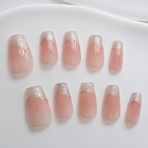 May include: A set of ten artificial nails. Each nail is a soft pink with a glittery silver tip and a thin gold line separating the two colors. The nails are oval-shaped and vary slightly in size.