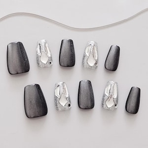 May include: A set of ten artificial nails. The nails alternate between a solid, gradient gray color and a silver glitter design with a reflective, abstract shape. The nails are square-shaped.