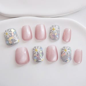 May include: A set of ten artificial nails in a light pink hue. Some nails feature a glittery silver base with white daisy accents and colorful sequins. The other nails are a solid, glossy pink.
