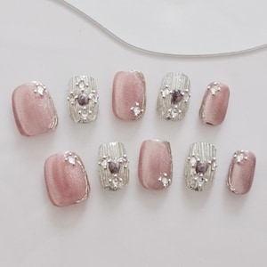 May include: A set of ten artificial nails. The nails alternate between a shimmering pink and silver design. The pink nails have a subtle sparkle, while the silver nails feature a textured pattern with small, decorative gems.