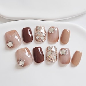 May include: A set of ten artificial nails in various shades of brown and beige. Some nails feature glitter, star-shaped embellishments, or a solid color. The nails are arranged on a white surface.