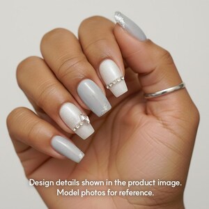 May include: Close-up of a hand with long, square-shaped artificial nails. The nails are painted in shades of white and gray, with glitter accents and rhinestone embellishments. The text at the bottom reads: Design details shown in the product image. Model photos for reference.