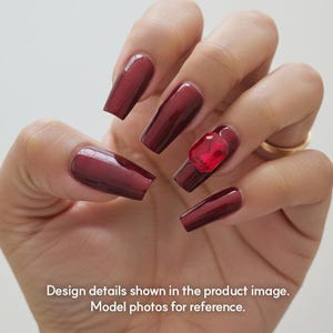 May include: A hand displaying long, burgundy-coloured artificial nails. One nail is adorned with a large, red, square-shaped gem. The nails have a glossy, solid finish. Text at the bottom reads: Design details shown in the product image. Model photos for reference.