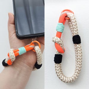 May include: A phone wrist strap with a woven rope design. The strap features orange, light blue, black, and beige colors. The strap is attached to a phone case with an orange ring.