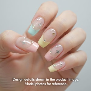 May include: A hand displaying a set of pastel-colored artificial nails. Each nail features a different design, including a light blue, yellow, and pink base with silver star and gem accents. The text at the bottom reads: Design details shown in the product image. Model photos for reference.