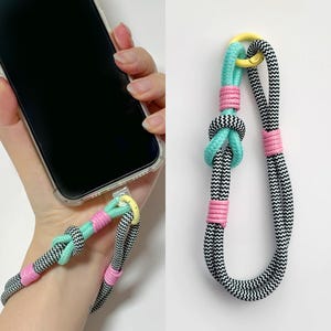 May include: A black and white braided phone strap with pink and turquoise accents. The strap has a yellow metal ring at the top and is tied in a knot at the bottom.