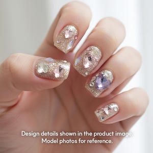 May include: Close-up of a hand with manicured nails. The nails are painted with a glittery gold polish and adorned with various sizes and shapes of rhinestones in pink, clear, and purple. The text at the bottom reads: Design details shown in the product image. Model photos for reference.