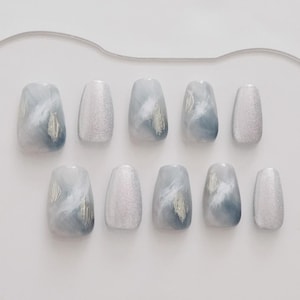 May include: A set of ten artificial nails in various shapes. The nails feature a design with a blend of light blue, white, and silver, creating a marbled effect. Some nails have a silver glitter finish.