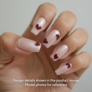 May include: Close-up of a hand with long, square-shaped nails. The nails are painted with a light pink, pearlescent polish and feature small, dark red heart designs. The text at the bottom reads: Design details shown in the product image. Model photos for reference.