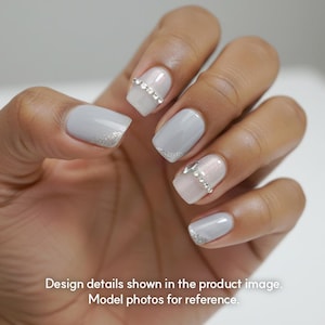 May include: A hand with manicured nails. The nails are painted in a light gray color with silver glitter accents on the tips and a line of rhinestones. The nails are square-shaped.