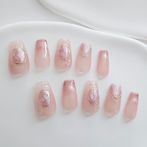 May include: A set of ten artificial nails in various shapes and sizes. The nails are primarily a light pink with a gradient effect. Some nails feature a metallic gold design and a glittery pink accent.