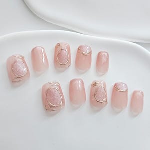 May include: A set of ten artificial nails in a light pink hue. Some nails feature a gold-colored outline with a glittery, oval-shaped design. The nails are arranged on a white surface, showcasing a modern nail art style.