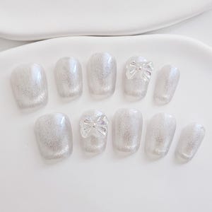 May include: A set of ten artificial nails in a light silver glitter finish. Two nails feature a clear bow accent. The nails are oval-shaped and arranged on a white surface, showcasing a delicate and elegant design.