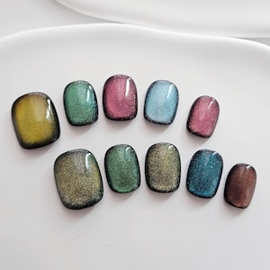 May include: A collection of ten oval-shaped artificial nails in various colors. The colors include yellow, green, pink, blue, and brown, each with a shimmering, glittery effect. The nails are arranged on a white surface.