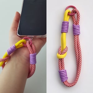 May include: A phone strap made of red, yellow, and purple rope with a pink metal ring. The strap is tied in a knot and has a loop for attaching to a phone.