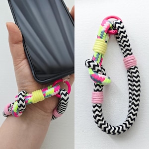 Puede incluir: Una colorida correa de muñeca para teléfono con un diseño rosa, negro, blanco y verde neón. La correa está unida a una funda de teléfono con un anillo rosa. La correa está hecha de cuerda trenzada.