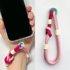 Puede incluir: Una correa para teléfono rosa y roja con detalles blancos y un cierre turquesa. La correa está hecha de cuerda trenzada y se puede usar para sujetar un teléfono a la muñeca o al dedo.