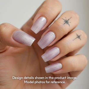 May include: Close-up of a hand with long, square-shaped nails painted in a shimmering, light purple shade. The nails have a glittery finish. The hand also has two small star tattoos on the fingers. The text at the bottom says "Design details shown in the product image. Model photos for reference."