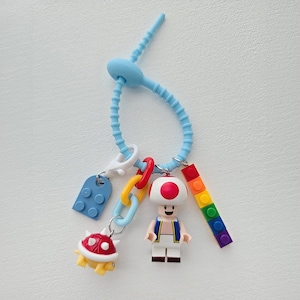 Puede incluir: Un llavero de silicona azul con varios dijes. Los dijes incluyen un ladrillo Lego azul, una tortuga roja y amarilla, una figura de Lego de Toad y un ladrillo Lego arcoíris. El llavero está sujeto a un fondo blanco.