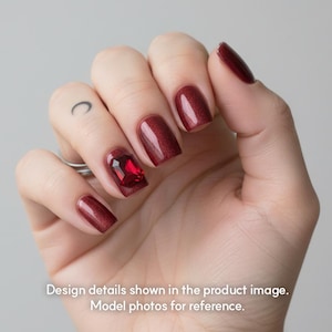 Peut inclure: Une main avec des ongles vernis d'une couleur rouge profond, certains avec un léger scintillement. Un ongle présente une grosse pierre précieuse rouge rectangulaire. Le texte de l'image est : "Design details shown in the product image. Model photos for reference."