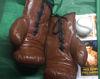 May include: A pair of brown leather boxing gloves with black laces. The gloves are tied together and are sitting on a green surface. The gloves have red stitching around the edges.