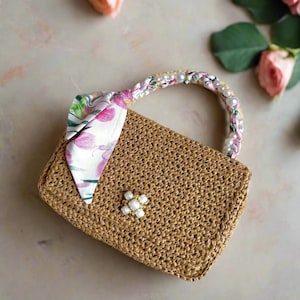 May include: A woven straw handbag with a rectangular shape. The handle is adorned with pearls and a floral fabric. A floral scarf is tied to the side, and a pearl flower embellishes the front. Pink roses are in the background.