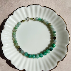 May include: A bracelet made of green beads, displayed on a white plate with a gold edge. The beads are various shades of green, from pale to dark, with some appearing almost black. The bracelet is secured with a silver clasp.