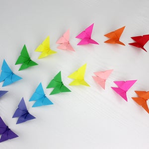 May include: A rainbow of origami butterflies arranged in a curved line on a white surface. The butterflies are made of paper in various colours, including red, orange, yellow, green, blue, purple, and pink.