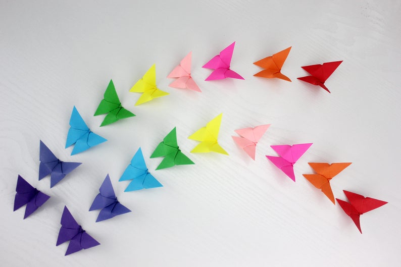 May include: A rainbow of origami butterflies arranged in a curved line on a white surface. The butterflies are made of paper in various colours, including red, orange, yellow, green, blue, purple, and pink.