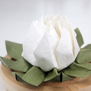 May include: A white origami lotus flower with green leaves sits on a wooden base.