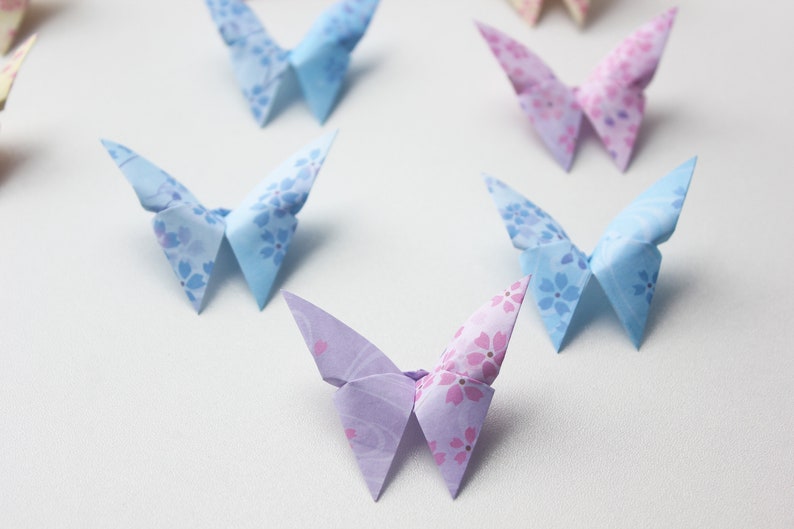 May include: A collection of origami butterflies with floral patterns in shades of blue, pink, and purple. The butterflies are arranged on a white surface.