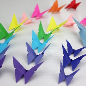 May include: A rainbow of origami butterflies, folded from paper in various colours, including purple, blue, green, yellow, pink, and red. The butterflies are arranged in a row on a white surface.