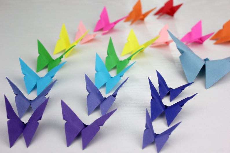May include: A rainbow of origami butterflies, folded from paper in various colours, including purple, blue, green, yellow, pink, and red. The butterflies are arranged in a row on a white surface.