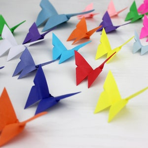 May include: A collection of colourful origami butterflies in various shades of blue, green, red, orange, yellow, purple, and white. The butterflies are arranged in a row on a white surface.