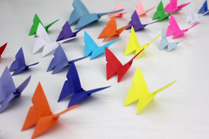 May include: A collection of colourful origami butterflies in various shades of blue, green, red, orange, yellow, purple, and white. The butterflies are arranged in a row on a white surface.