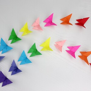 May include: A rainbow of origami butterflies arranged in a semi-circle on a white surface. The butterflies are made of paper in various colours, including red, orange, yellow, green, blue, indigo, and violet.