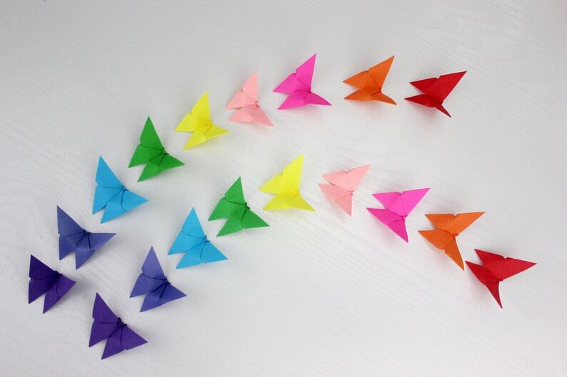 May include: A rainbow of origami butterflies arranged in a semi-circle on a white surface. The butterflies are made of paper in various colours, including red, orange, yellow, green, blue, indigo, and violet.