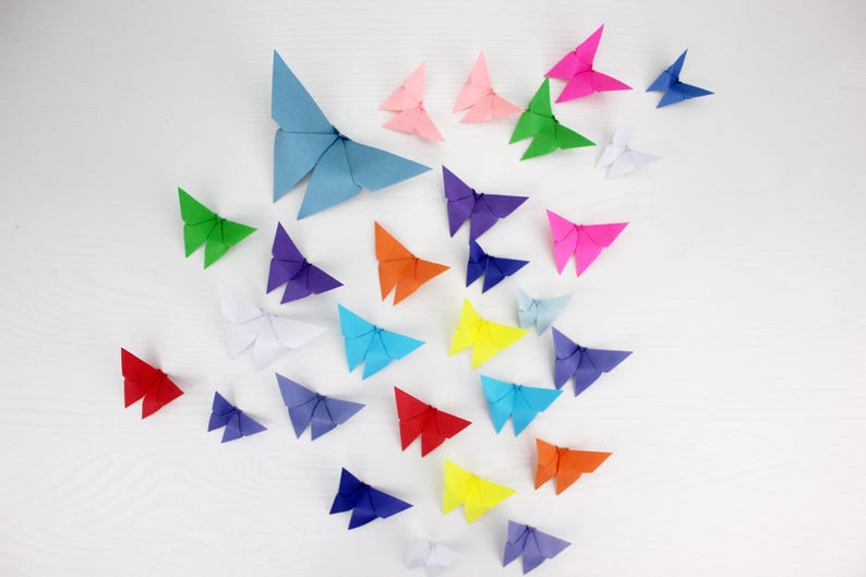 May include: A collection of colourful origami butterflies in various shades of blue, green, red, pink, orange, yellow, and purple. The butterflies are arranged in a cluster on a white surface.