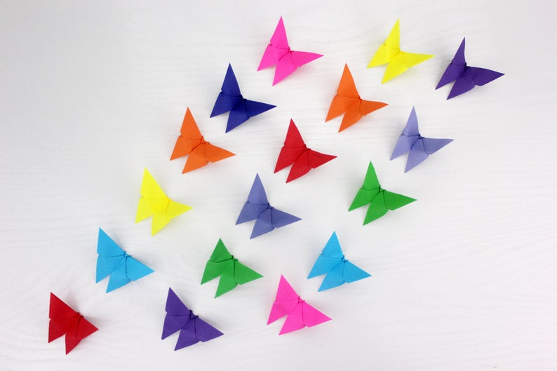 May include: A collection of 17 origami butterflies in a rainbow of colours, including red, orange, yellow, green, blue, purple, and pink. The butterflies are arranged in a loose pattern on a white surface.