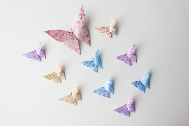 May include: A collection of ten origami butterflies in various pastel colors, including pink, blue, and yellow, with floral patterns. The butterflies are arranged on a white background.